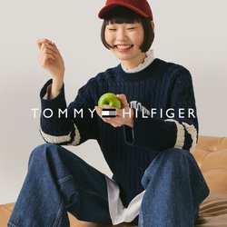 Mladá osoba v červené čepici, tmavě modrém svetru a džínách sedí na krémově hnědé kožené pohovce, drží zelené jablko a usmívá se na jednobarevném pozadí.