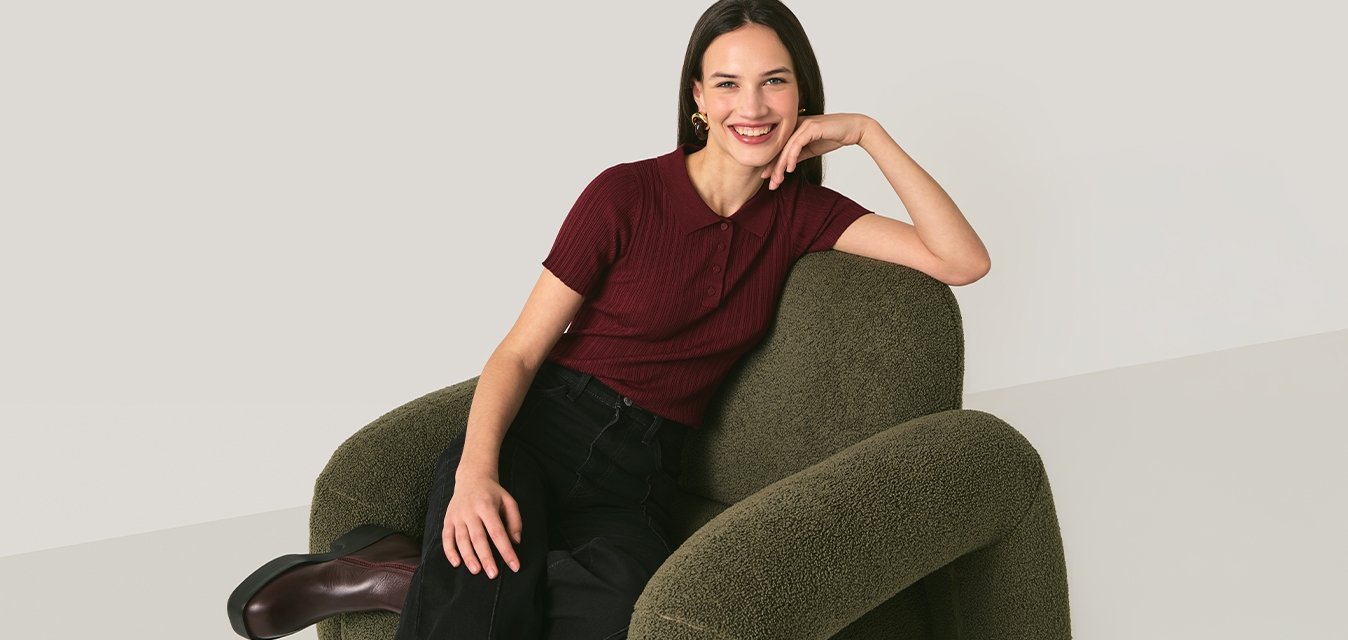 Mladá žena se usmívá, sedí na zeleném texturovaném křesle, má na sobě vínovou košili, černé kalhoty a hnědé boty, na jednobarevném pozadí.