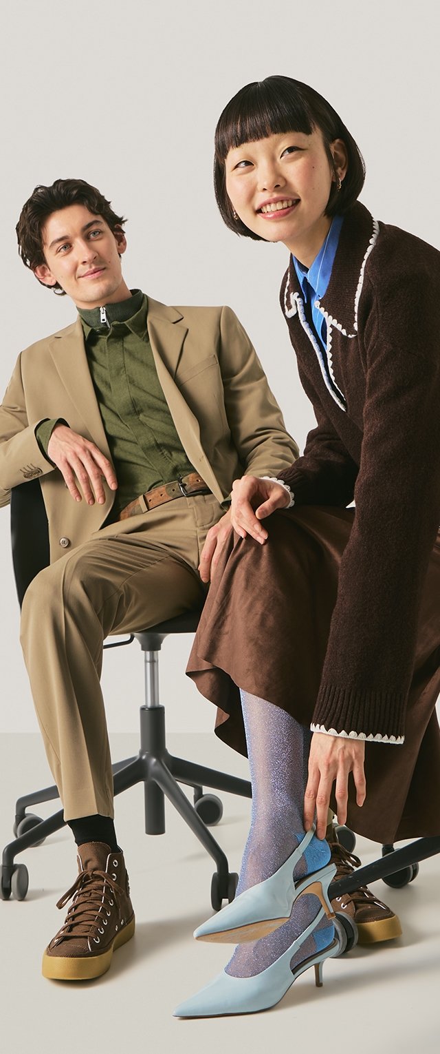 Mujer sonriente con atuendo marrón se pone tacones azules con medias brillantes, hombre con traje beige y camisa verde sentado en una silla de oficina cerca.