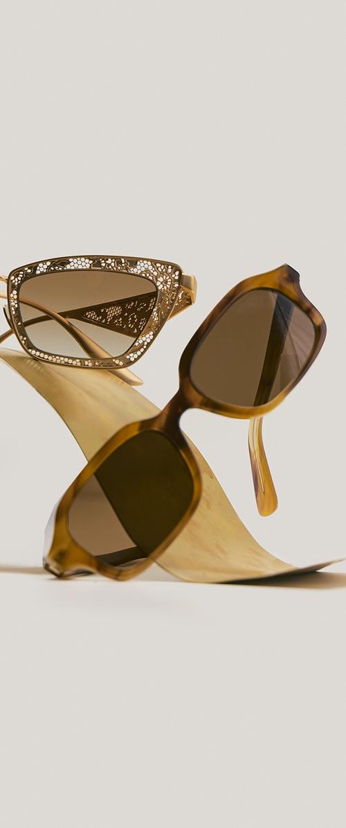 Deux paires de lunettes de soleil marron, l'une avec des montures en filigrane doré ornées et l'autre avec des montures rectangulaires en écaille de tortue, expos ées sur une surface dorée incurvée.