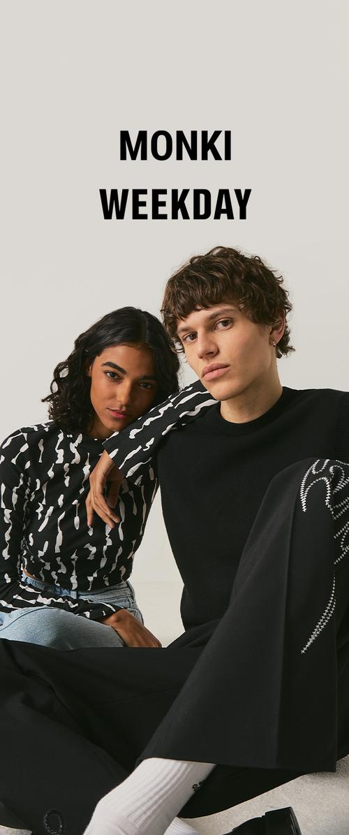 Dos modelos posan sentados; la mujer lleva una blusa negra con estampado y vaqueros, el hombre lleva un suéter negro y pantalones con calcetines blancos; texto "MONKI WEEKDAY" arriba.