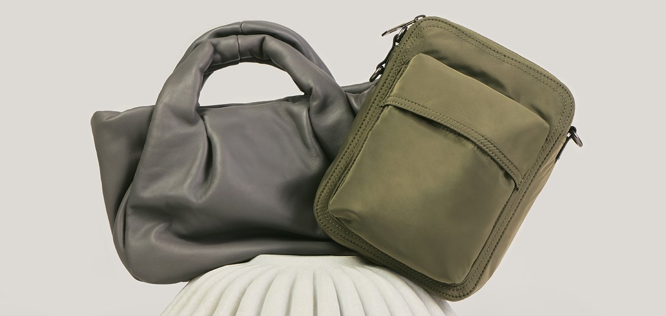 Dvě kabelky, jedna šedá měkká kožená tote taška a jedna olivově zelená obdélníková crossbody taška, vystavené na bílém texturovaném stojanu.
