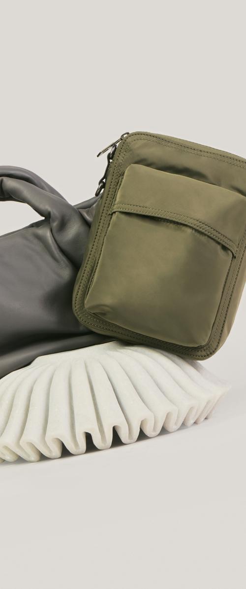 Olivgrüne, mit Reißverschluss versehene Tasche mit Vordertasche, die auf einem schwarzen Lederhandschuh und einer weißen, plissierten Stoffoberfläche liegt.