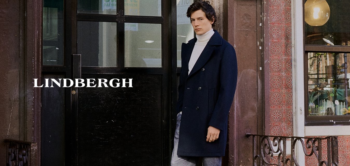 Muž v námořnickém kabátu a šedých kalhotách stojí na betonových schodech před cihlovou budovou s velkými okny a černými dveřmi.