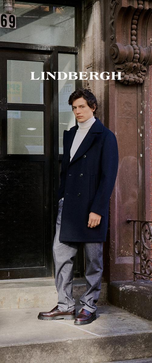 Abrigo azul marino de doble botonadura con cuello, suéter de cuello alto crema, pantalones grises y mocasines marrones con una textura distintiva, de pie sobre escalones de piedra.