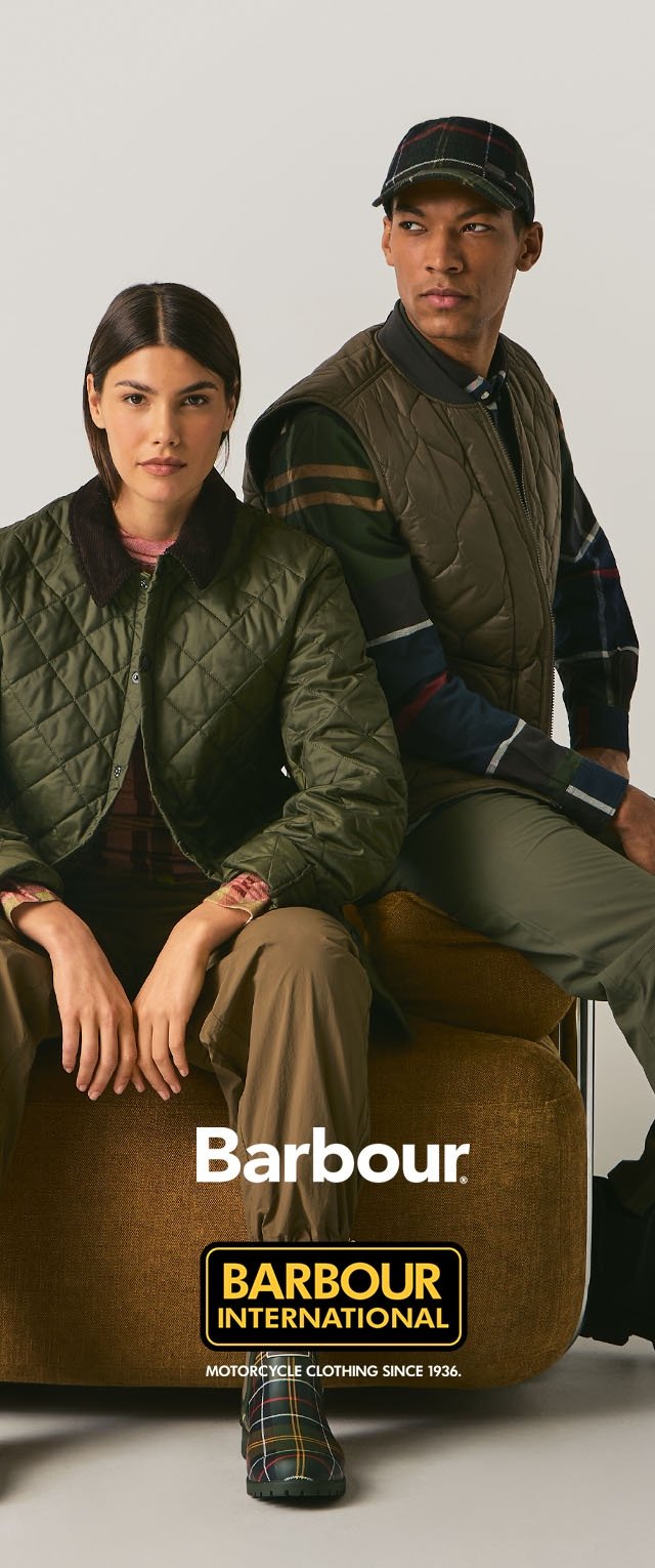 Woman and man seated on brown chair wearing green quilted jackets and plaid patterns, promoting Barbour International motorcycle clothing.