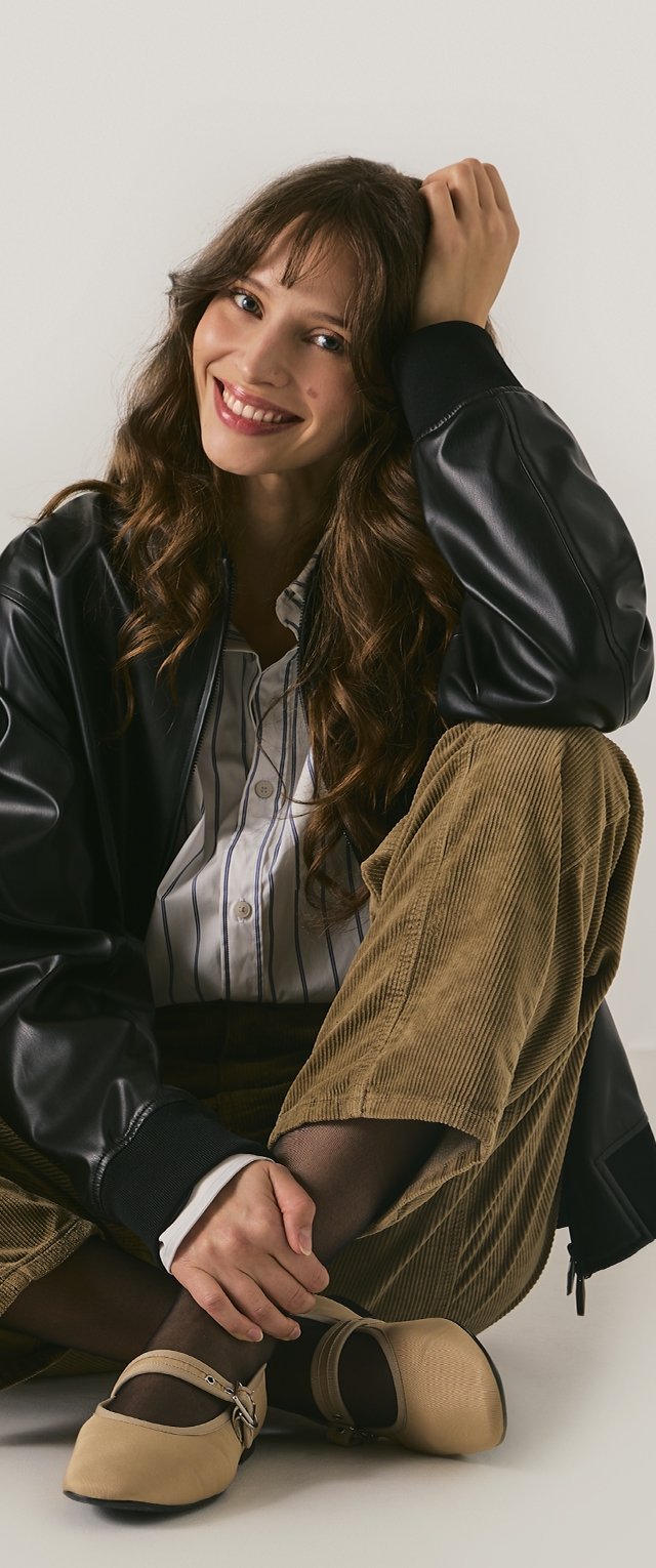 Una mujer está sentada con las piernas cruzadas, vistiendo una chaqueta bomber de cuero negra, una camisa de rayas blanca y pantalones de pana marrones con bailarinas beige que tienen un hebilla.