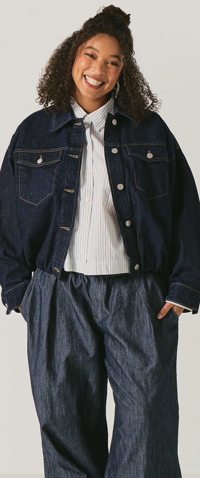 Mujer con cabello rizado sonriendo, vistiendo una chaqueta de mezclilla oscura de gran tamaño, camisa de rayas abotonada y pantalones holgados de mezclilla oscura, con las manos en los bolsillos.