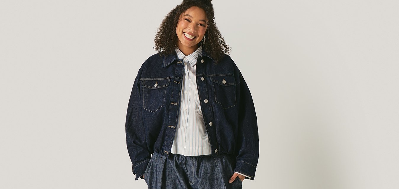 Usmívající se osoba stojící, oblečená do tmavé volné bundy, volných tmavých kalhot a bílé košile, na jednobarevném světlém pozadí.