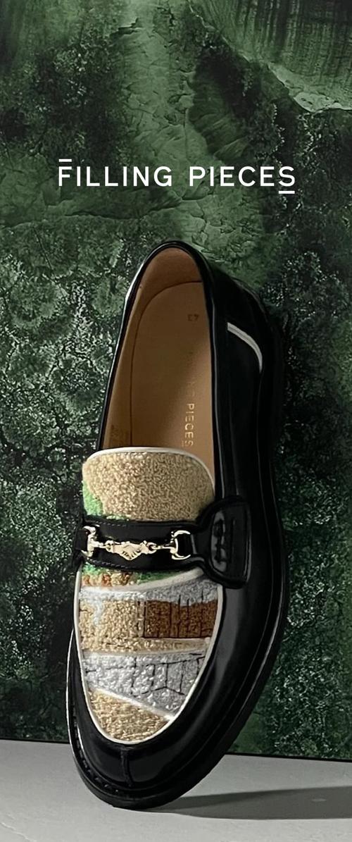 Mocasín negro con patchwork multicolor texturizado en la parte delantera y detalle de horsebit plateado sobre un fondo texturizado verde. El texto dice "Filling Pieces."