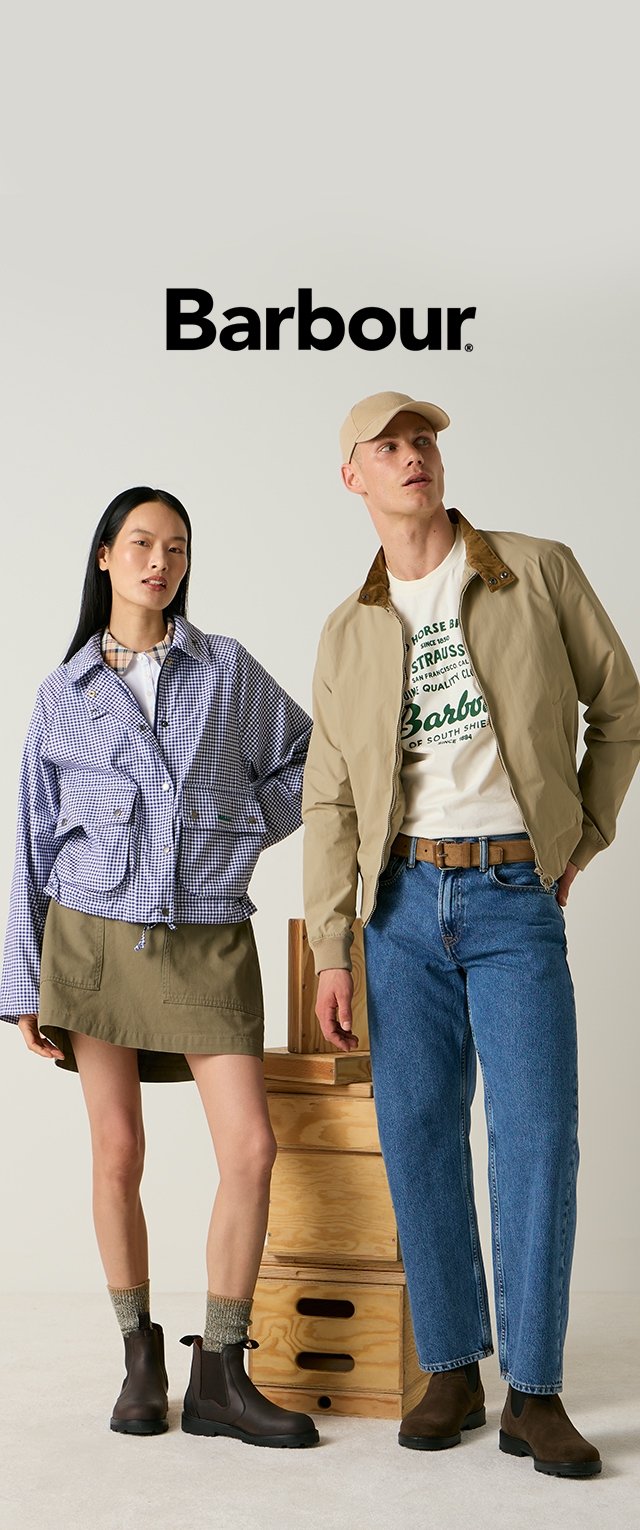 Junge Frau und Mann tragen lässige Barbour-Outfits, darunter Jacke, Rock, Jeans, Stiefel und Mütze, und stehen neben Holzkisten vor einfarbigem Hintergrund.