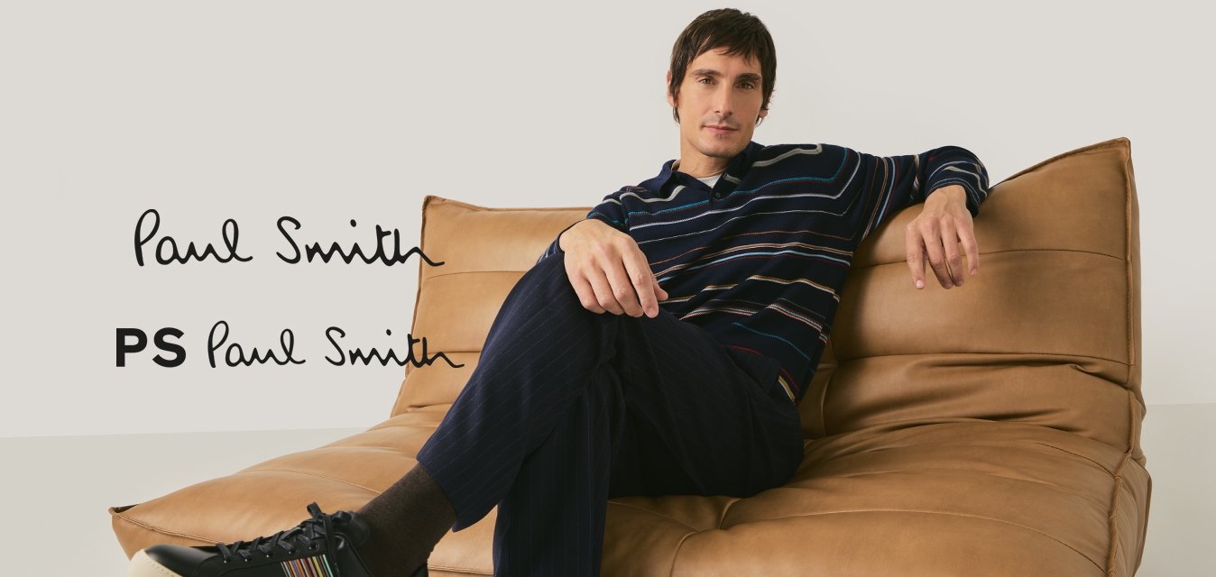 Muž v proužkované košili s dlouhým rukávem a tmavých kalhotách sedící s překříženýma nohama na hnědém koženém gauči na neutrálním pozadí.