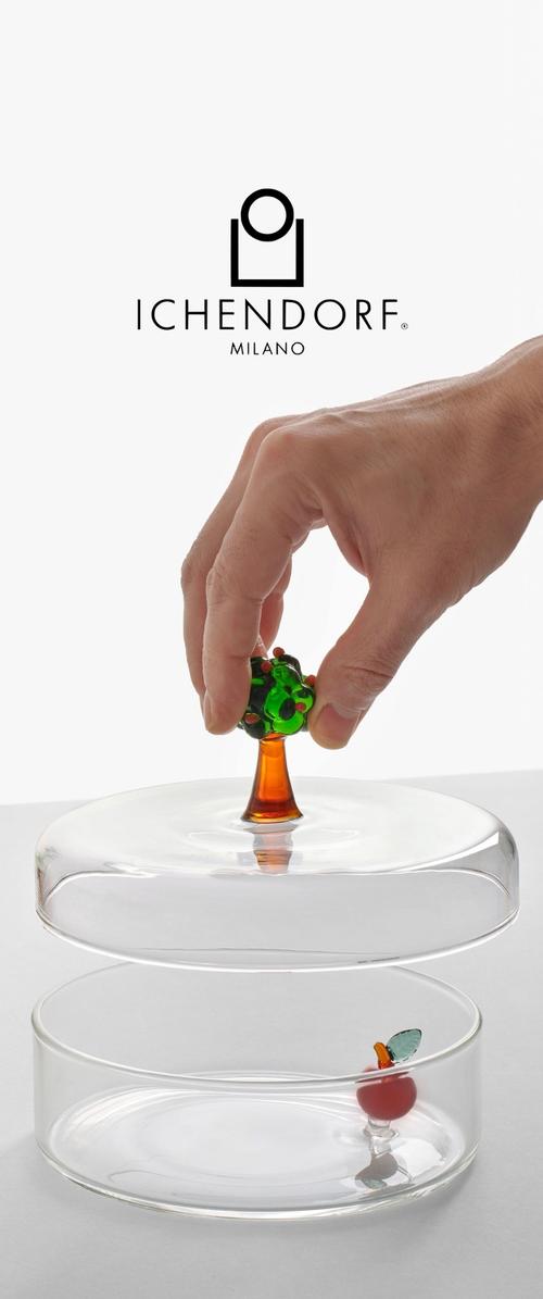 Mano levantando una cúpula de vidrio con un asa en forma de árbol, revelando una pequeña manzana de vidrio dentro de un recipiente redondo y transparente, con el logo de Ichendorf Milano arriba.