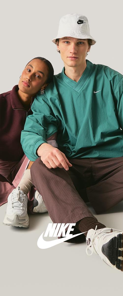 Dwoje młodych dorosłych siedzi blisko siebie; jedna osoba ma na sobie zielony top Nike i biały kapelusz typu bucket, druga granatową kurtkę, oboje w butach Nike na jasnym tle.