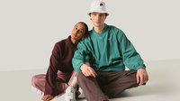 Zwei Personen sitzen auf dem Boden, eine Frau in einer burgunderfarbenen Jacke und pinken Hosen lehnt sich an einen Mann in einer grünen Nike-Jacke, braunen Hosen und weißem Anglerhut.