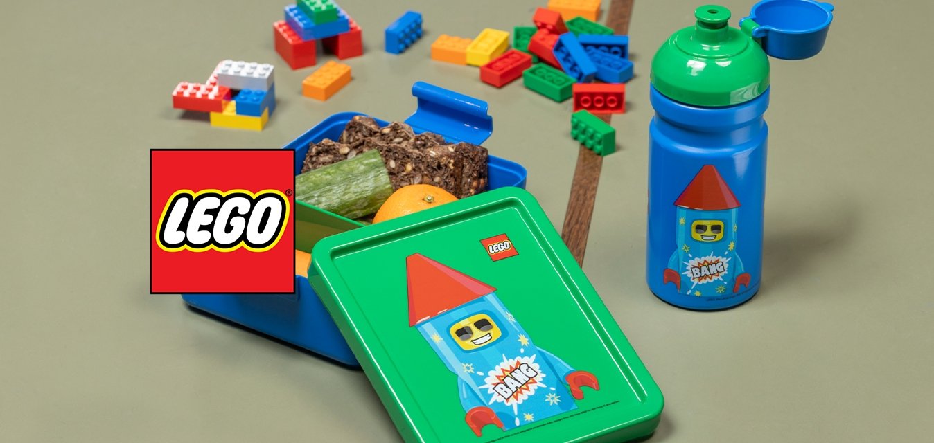 Modrá svačinová krabička s tématem LEGO se zeleným víkem, plná svačinek a pomeranče, vedle odpovídající modré lahve se zeleným uzávěrem, na stole.
