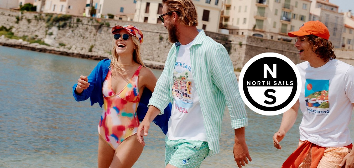 Tři přátelé v barevném letním oblečení se procházejí a smějí se u vodního toku, v pozadí jsou historické budovy a nad nimi jasná obloha.