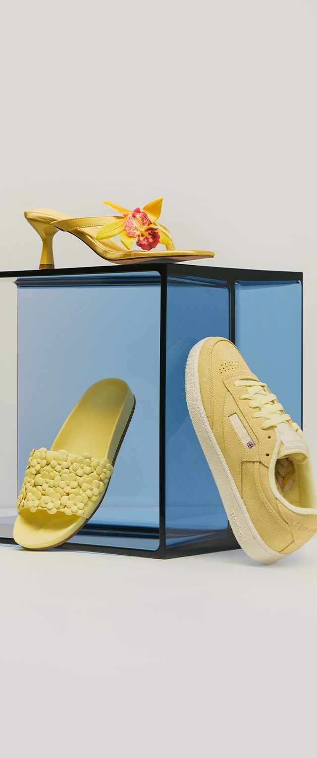 Tres zapatos amarillos: una chancla floral con adornos de flores en 3D, una sandalia de tacón con una orquídea amarilla y roja, y una zapatilla de ante sobre un cubo azul.