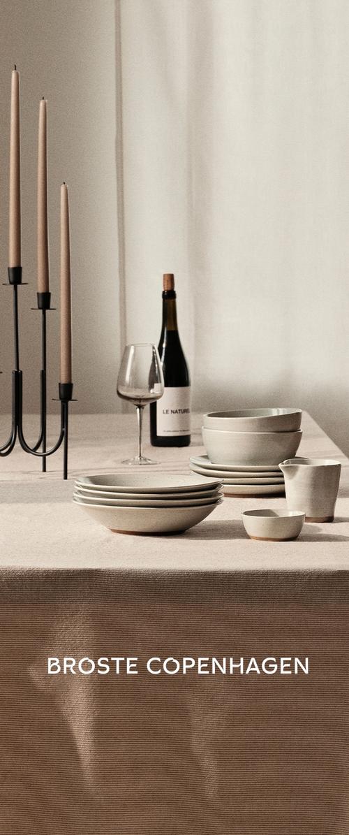 Mesa de comedor minimalista con velas beige en candelabros negros, platos blancos de cerámica apilados, cuencos, una jarra para crema, copa de vino y botella de vino.