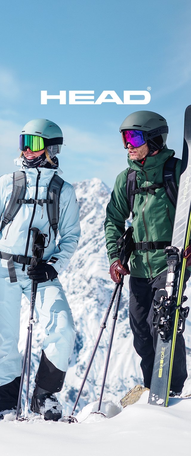 Esquiadores con chaquetas de invierno azul claro y verde oscuro están de pie en la nieve. Ambos llevan cascos y gafas, sosteniendo bastones de esquí y esquís. Fondo montañoso.