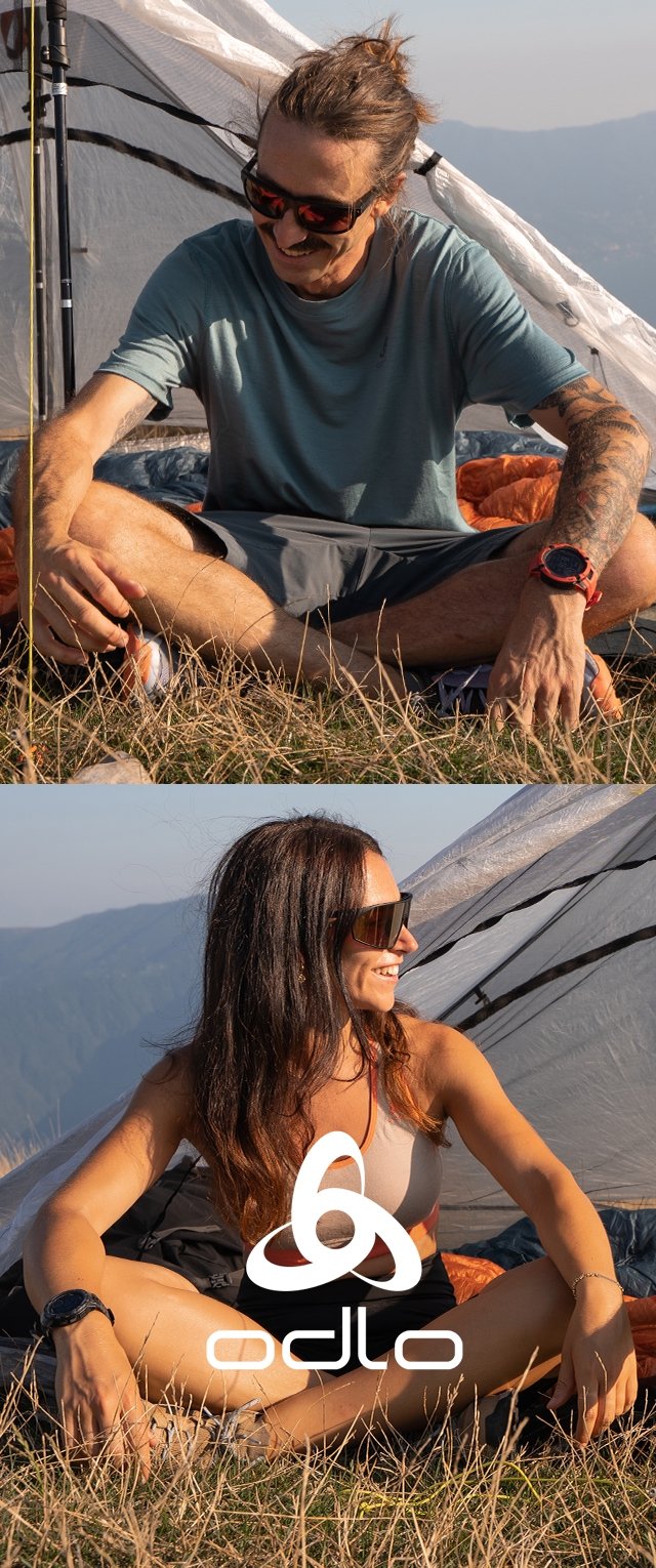 Man en vrouw met zonnebrillen en sportkleding zitten met gekruiste benen voor een tent op grasland, glimlachend in de buitenlucht bij zonsondergang.