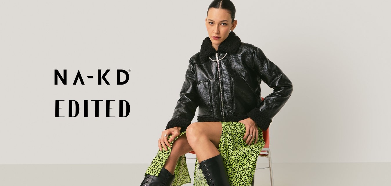 Žena sedící na židli, oblečená v černé kožené bundě, neonově zelené rozdělené sukni a vysokých černých botách na šněrování na jednobarevném pozadí.