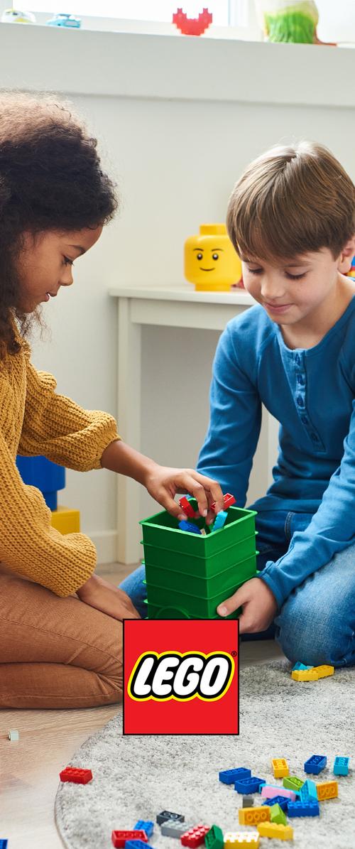 Deux enfants sont assis par terre et jouent avec des briques LEGO provenant d’une boîte verte, avec des pièces éparpillées et un logo LEGO au premier plan.