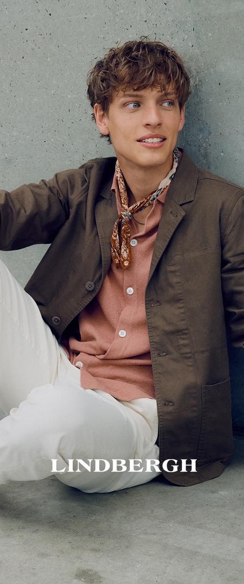 Blazer marrón ligero sobre una camisa de botones rosa, acompañado de pantalones blancos y una bufanda con estampado, sentado contra una pared gris.