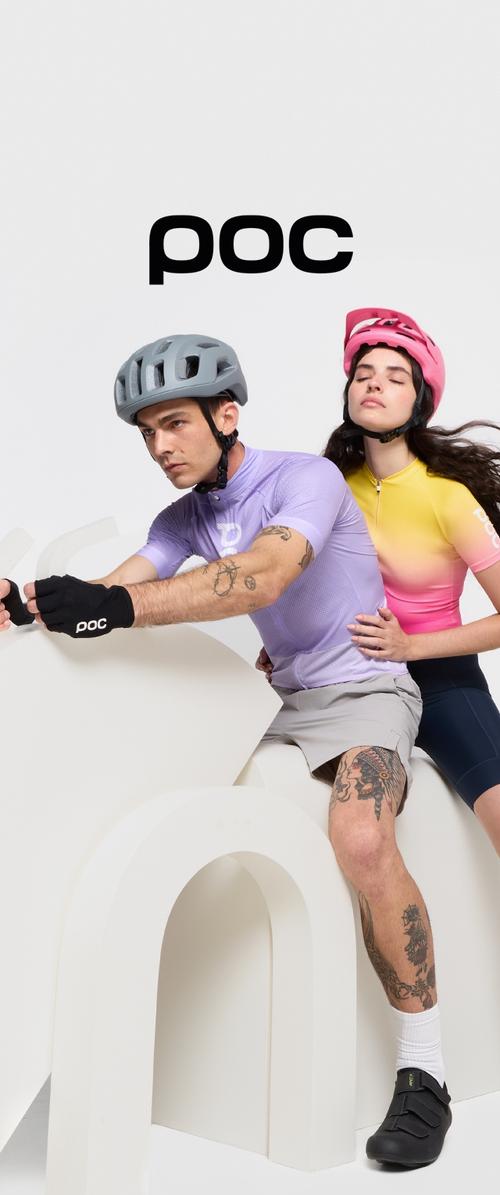 Two cyclists wearing performance gear pose on a curved white structure. He has a light purple shirt, grey shorts, and a grey helmet; she wears a pink and yellow gradient top.