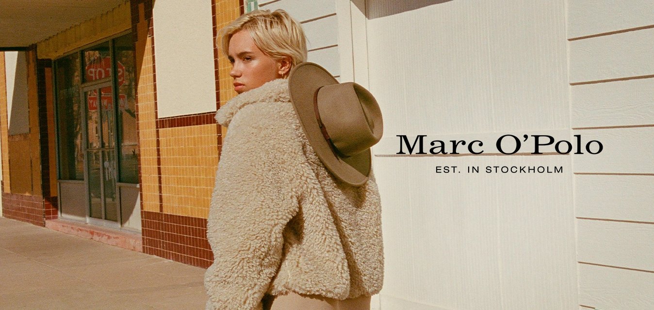 Béžová chlupatá bunda z ovčí vlny s vysokým límcem, nošená přes krémový svetr. Hnědé kalhoty, zelený klobouk se širokou krempou položený na bundě.