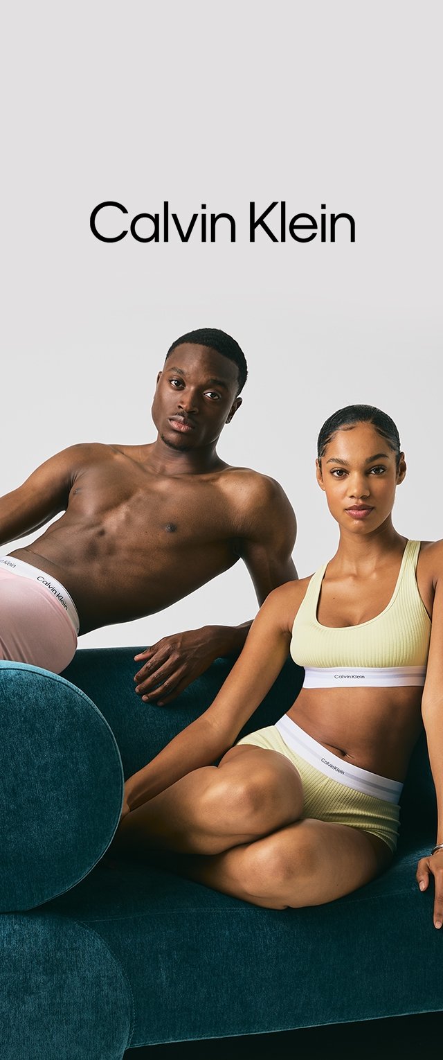 Dos modelos vestidos con ropa interior de Calvin Klein están sentados en un sofá color teal frente a un fondo sencillo con el logotipo de Calvin Klein arriba.