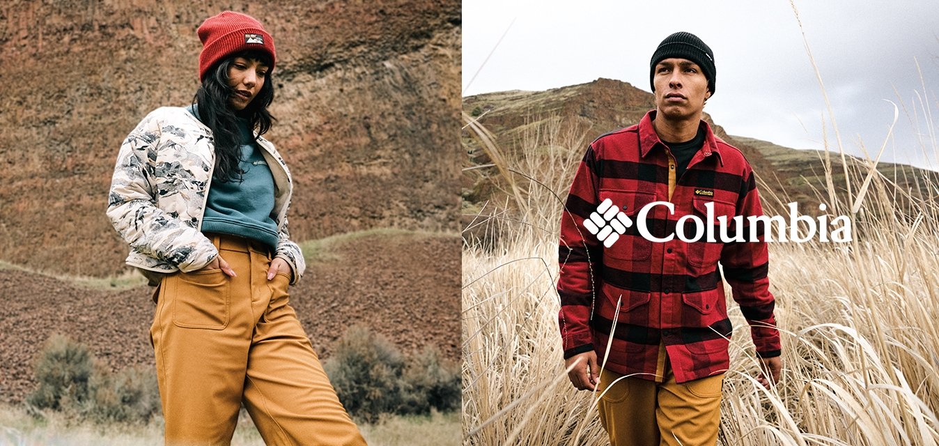 Mladá žena s červenou čepicí, květinovou bundou, tyrkysovým topem a hořčicovými kalhotami stojí na skalnatém terénu; muž v černé čepici a červené kostkované bundě ve vyschlé trávě.