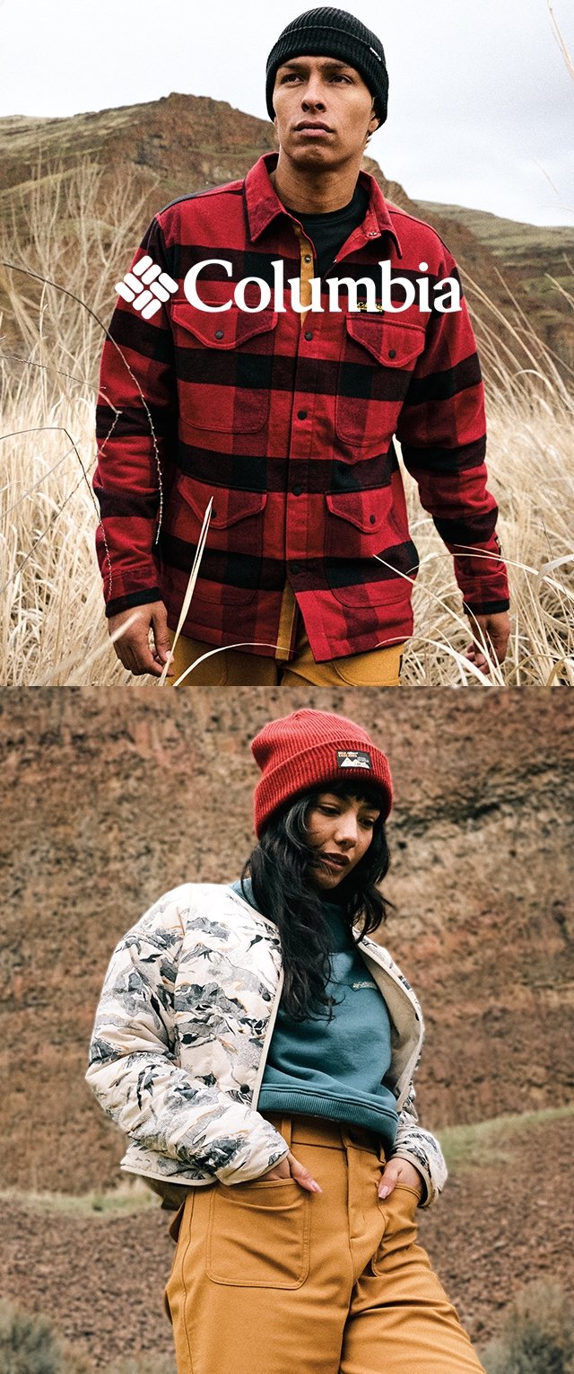 Hombre con chaqueta de cuadros roja y negra y gorro negro en hierba alta; mujer con chaqueta floral, camiseta turquesa, gorro rojo y pantalones beige cerca de terreno rocoso.