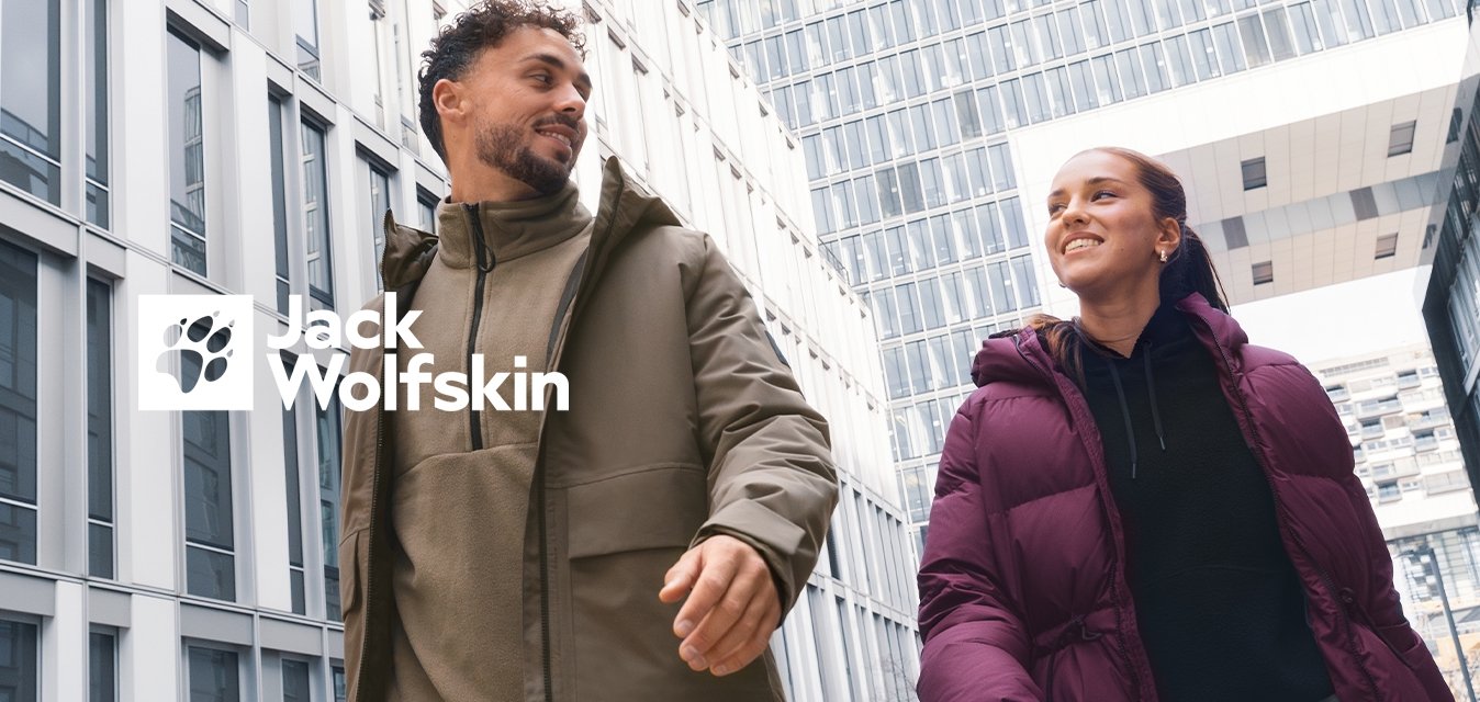 Dva lidé v zimních bundách kráčejí a usmívají se před moderními kancelářskými budovami v městském prostředí za zataženého dne.