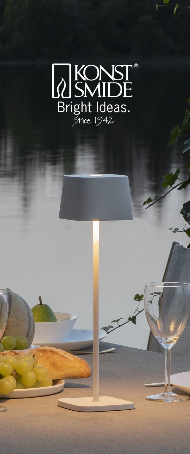 Lámpara de mesa blanca con pantalla cilíndrica y base plana, iluminando suavemente. Colocada sobre una mesa gris con uvas y pan cerca.