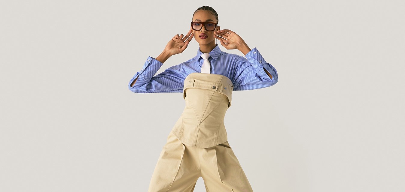 Model je na fotografii v béžových širokých kalhotách a korzetu přes modrou pruhovanou košili s bílou kravatou, doplněná brýlemi a náušnicemi.