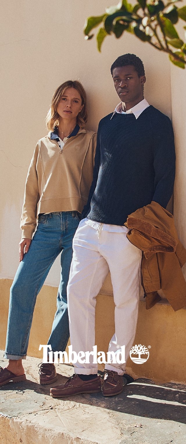 Two individuals stand against a wall. One wears a beige quarter-zip sweatshirt, blue jeans, and brown shoes. The other wears a navy ribbed sweater, white pants, and brown shoes.