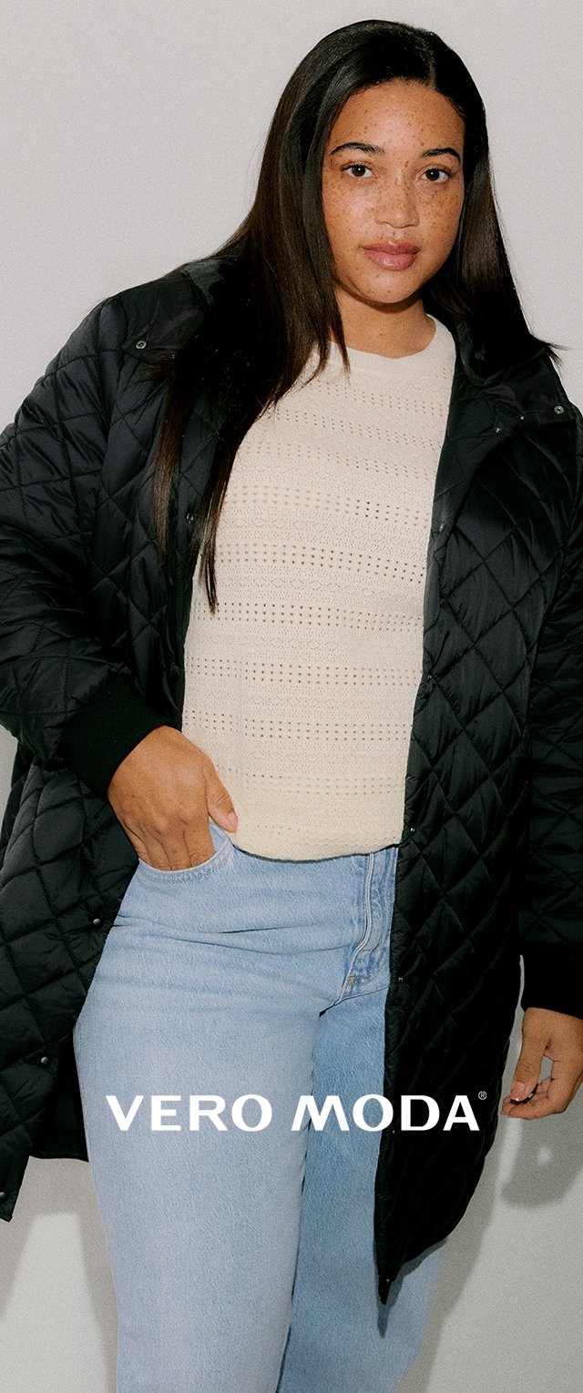 Zwarte gewatteerde jas met ritssluiting, voorzien van geribbelde manchetten, gedragen over een licht gebreide trui en lichtblauwe spijkerbroek.