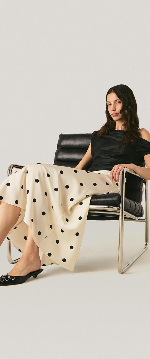 Mujer con cabello largo y oscuro sentada en una silla de cuero negra, vestida con un top negro, falda crema con lunares negros y zapatos negros de tacón bajo.