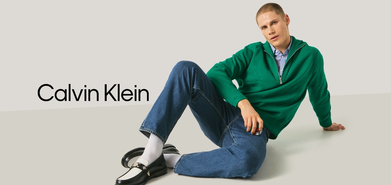 Mladý muž ležící na podlaze, oblečený v zeleném svetru, modrých džínách, bílých ponožkách a černobílých mokasínách, držící Rubikovu kostku.