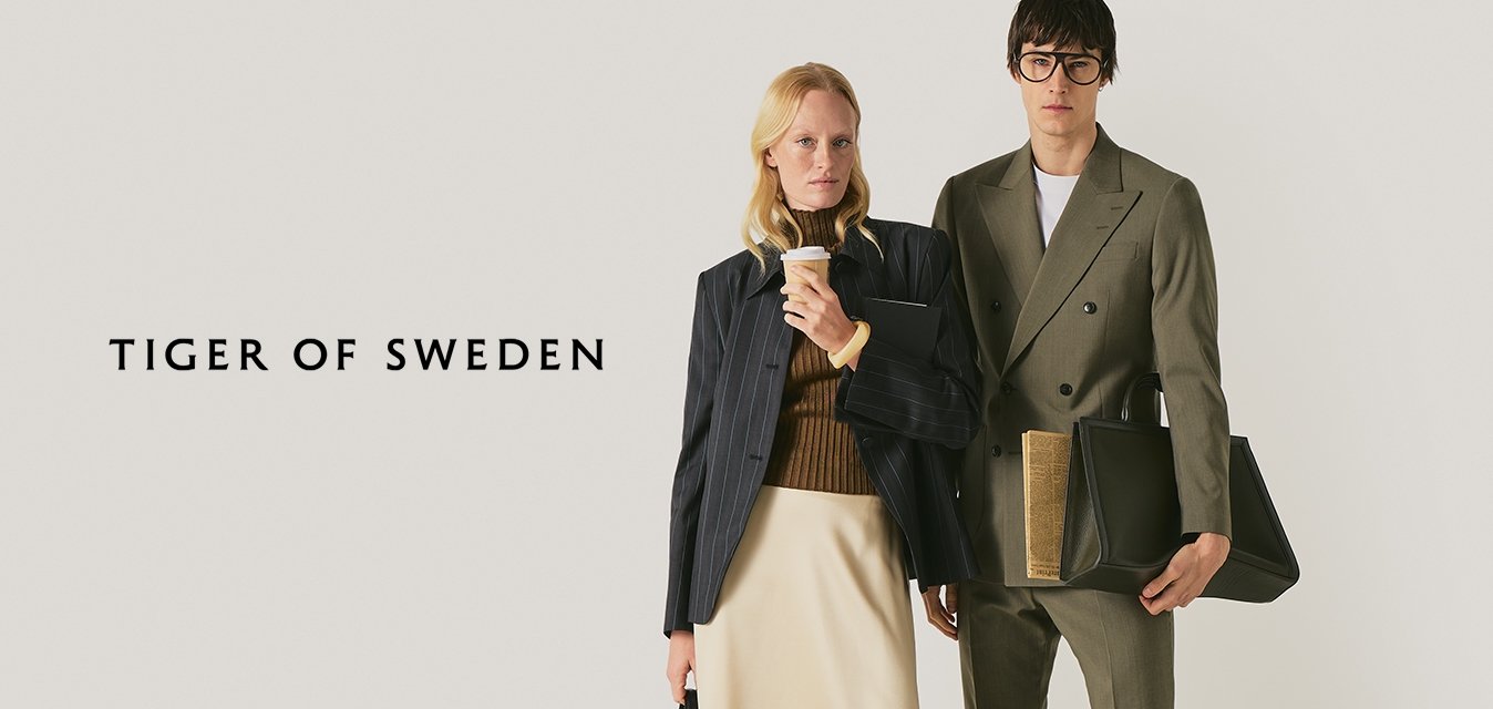 Dva profesionálně oblečení lidé stojí: žena v béžové sukni, černých botách, držící kávu a kabelku; muž v olivovém obleku držící velkou černou tašku.