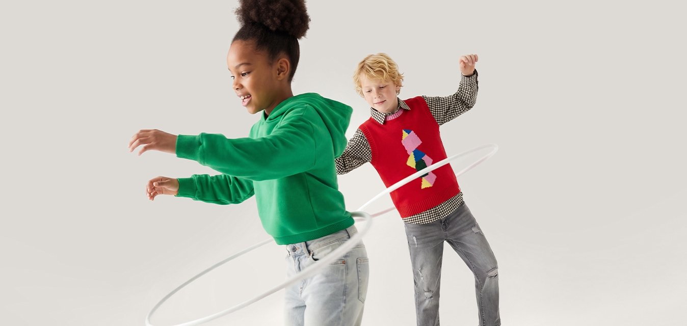 Dvě děti si hrají s hula hoopem, jedno ho točí kolem pasu, zatímco druhé sleduje a usmívá se.