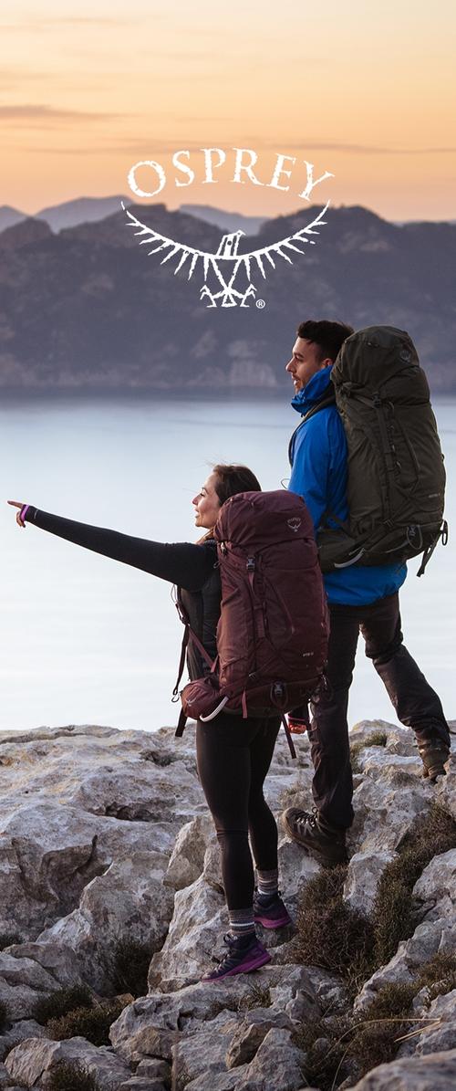 Dos excursionistas con mochilas grandes están de pie sobre un terreno rocoso al atardecer, uno señalando hacia las montañas distantes sobre un lago en calma.
