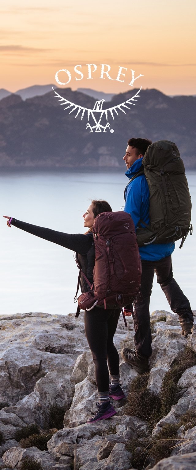 Zwei Wanderer mit großen Rucksäcken stehen auf felsigem Gelände bei Sonnenuntergang, einer deutet auf die entfernten Berge über einem ruhigen See.