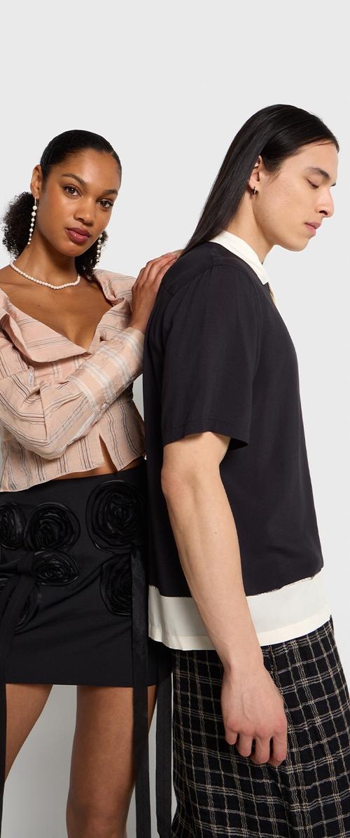 Dos individuos visten atuendos contrastantes: uno lleva una blusa corta beige a rayas y una falda negra con detalle de rosa; el otro viste una camisa negra y pantalones a cuadros.