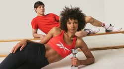Frau in rotem Sport-BH und schwarzen Leggings hält eine Hantel, Mann in rotem Shirt und Shorts lehnt an der Stange im Fitnessstudio.