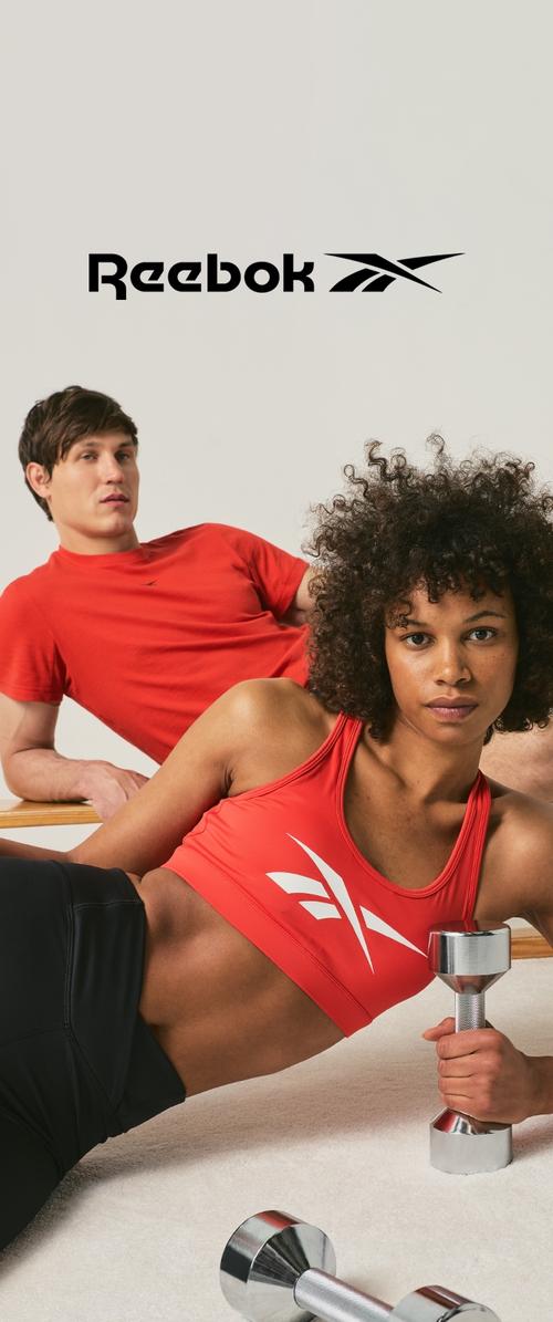 Žena v červeném sportovním topu Reebok drží činku, muž v červeném tričku Reebok za ní, logo Reebok nahoře na neutrálním pozadí.