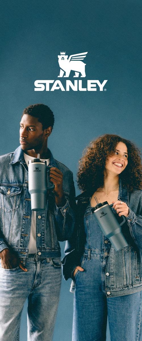 Dos personas con vaqueros sosteniendo vasos térmicos grises Stanley, de pie contra un fondo azul debajo del logo de Stanley.