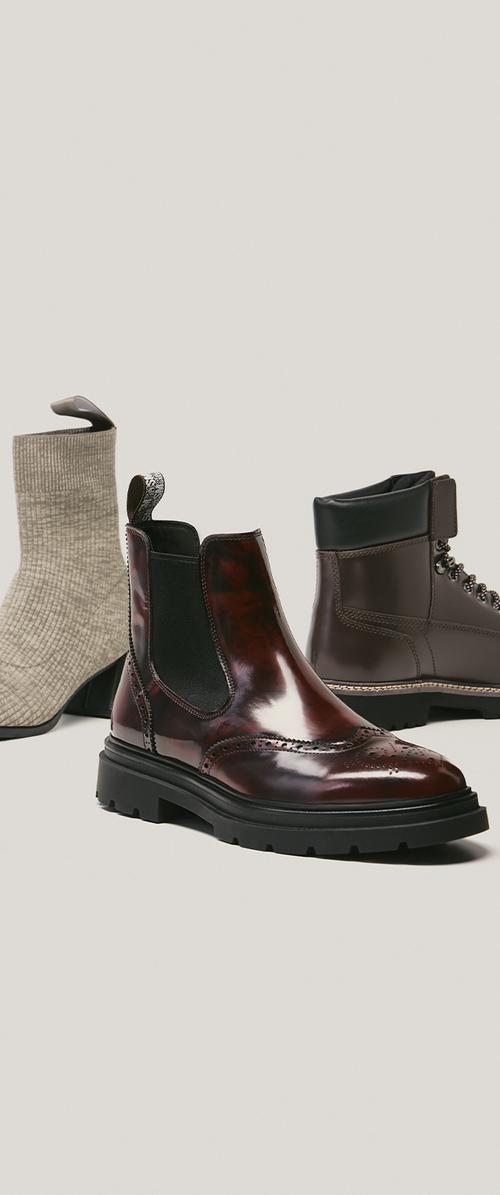 Three ankle boots: polished dark brown leather Chelsea boot, knitted beige sock-style boot, and matte brown lace-up boot with padded collar.