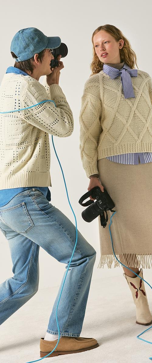 Photographer in jeans and knit sweater takes picture of woman wearing knit sweater, bow blouse, beige skirt, and cowboy boots.